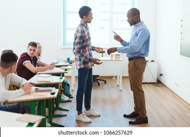 Side view of african american teacher pointing at wristwatch to latecomer schoolboy in classroom