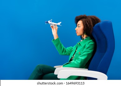 Side View Of African American Woman Holding Model Of Plane And Sitting On Seat Isolated On Blue