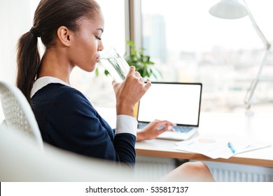 Side view of African business woman in dress sitting by the table in office and drinking water