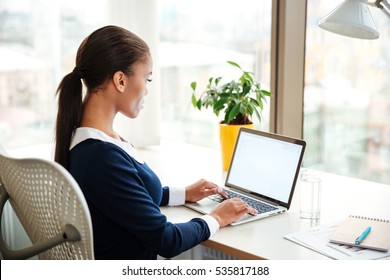 Side View Of African Business Woman In Dress Sitting On Armchair Near The Window And Using Laptop In Office