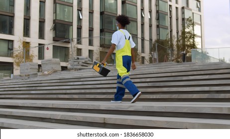 Side view of african construction worker in safety mask walking on street during reconstruction. Afro american repairman in uniform carrying toolkit working at construction site