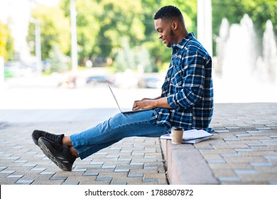 Side View Of African Guy Working On Laptop Sitting On The Border In Downrown, Selective Focus