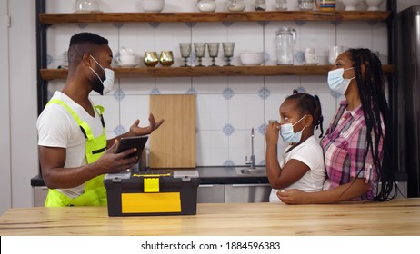 Side View Of African Plumber In Safety Mask Holding Digital Tablet Talking To Woman Client. Afro-american Female Customer With Daughter Wearing Protective Mask Talking To Handyman In Kitchen