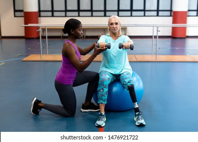 Side view of African-american Female trainer assisting disabled Caucasian senior woman to exercise with dumbbell in sports center. Sports Rehab Centre with physiotherapists and patients working - Powered by Shutterstock