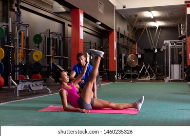 Side View Of An  African-American Woman Doing A Single Leg Raise While An African-American Man Assists Her Inside A Room At A Sports Center