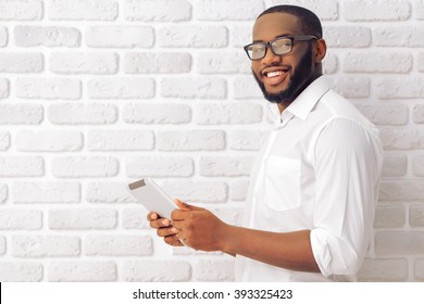 Side view of Afro American man in classic shirt and glasses using a tablet, looking at camera and smiling, standing against white brick wall