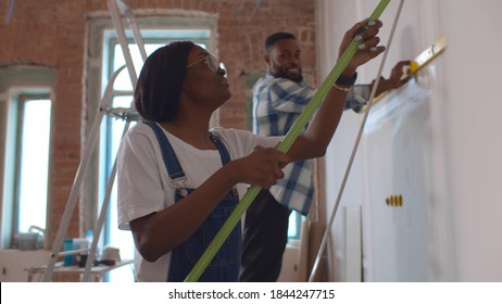 Side View Of Afro-american Couple Doing House Renovation Together. African Woman Painting Wall With Roller Brush While Her Boyfriend Using Measurement Level Redecorating Apartment