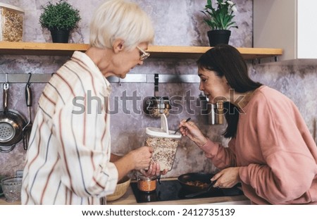 Side view of aged Asian woman scooping spices out of container with spoon while ethnic girlfriend helping cooking lunch in kitchen