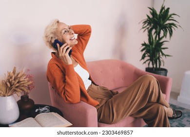 Side view of aged retired female in casual jacket having conversation on cellphone and smiling while sitting on couch at home - Powered by Shutterstock