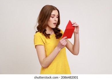 Side View Of Amazed Young Woman In Yellow Casual T-shirt Opening Envelope And Reading Letter, With Surprised Expression, Got Unexpected Pleasant News. Indoor Studio Shot Isolated On Gray Background.