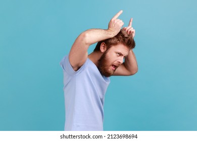 Side View Of Angry Bully Bearded Man Showing Bull Horn Gesture With Fingers Over Head, Looking Hostile And Threatening, Aggressive Face. Indoor Studio Shot Isolated On Blue Background.