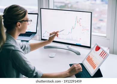 Side view anonymous young female analyst pointing and stylus at desktop computer while studying chart near tablet at work