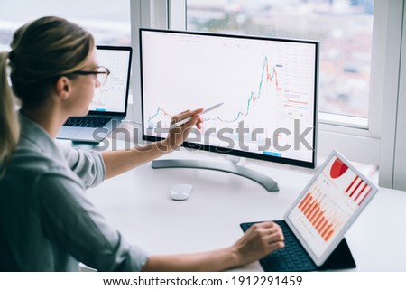 Side view of anonymous young female analyst pointing with stylus at desktop computer while studying chart near tablet at work