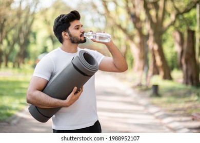 Side View Of Arabian Bearded Man Wearing Sport Clothes Drinking Fresh Water From Bottle While Standing At Green Park. Handsome Athlete Taking Break During Outdoors Workout.