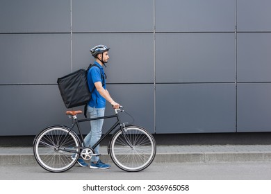 Side view of arabian courier with thermo backpack walking near bike and building outdoors