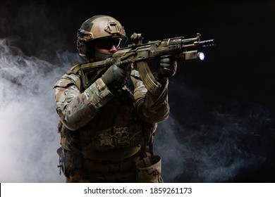 Side View Of Army Soldier In Military Camouflage Uniform, Helmet, Glasses And Mask Holds Rifle Near Face And Takes Aim Using Collimator Sight Standing In Attack Position, In Smoke On Black Background