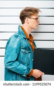 Side View Of Arrested Hipster Man In Eyeglasses Holding Empty Prison Board In Front Of Police Line Up