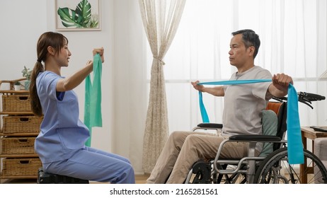 Side View Of Asian Disable Mature Man Sitting In Wheelchair And Working Out With Female Personal Physiotherapy Care Attendant’s Help. They Use Resistance Band Together At Home