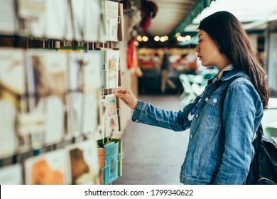 Side View Asian Female Exchange Student Is Selecting Postcards From A Rack. Selective Focus Woman Traveling Alone Is Picking Gift At The Exterior Of A Souvenir Store. Genuine Lifestyle