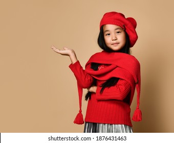 Side view of an Asian girl in a red hat, scarf and sweater, holding her palm and looking at the camera. Child posing on a beige background in the studio.