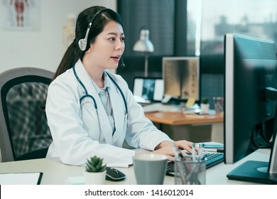 Side View Of Asian Japanese Woman Doctor Vet Sitting With Headset At Desk In Front Of Computer Talking Soothingly With Patient Telemedicine Concept. Female Medical Nurse Listening And Typing Keyboard