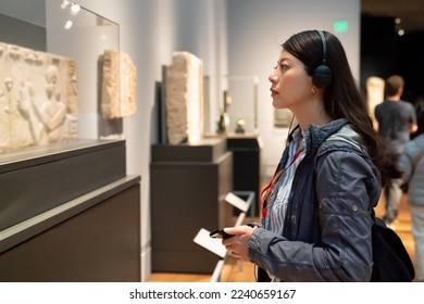 side view of asian Korean college girl taking audio tour in the history museum located in los angeles usa. she is looking at an ancient carving displayed in glass case
