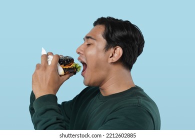 Side View. Asian Man Eating Burger Posing Standing Over Pastel Blue Studio Background. Black Guy Tasting Black Burger Enjoying Unhealthy Junk Food. Nutrition And Overeating Habit