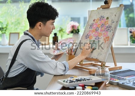 Side view of asian man painting with watercolor on canvas in bright living room. Art, creative hobby and leisure activity concept