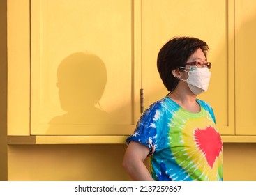 Side View Of Asian Middle Aged LGBT Woman In Rainbow T-shirt With Protective Mask In Thoughtful Expression While Standing Near Yellow Wooden Street Coffee Kiosk In Public Area