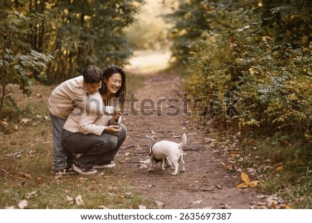 Side view of an Asian mother and her young son laughing while walking a small white dog on a forest trail. They wear casual autumn clothes. Great for family, parenting, and outdoor lifestyle themes.