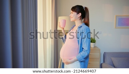 Side view of asian pregnant woman holding a cup of water standing near window at home with smiling