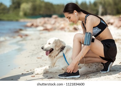 Side View Of Asian Sportswoman In Earphones With Smartphone In Running Armband Case Tying Shoelaces Near Golden Retriever On Beach 