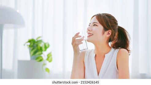 side view of asian woman with brunette ponytail smile drinks a glass water happily in a white room