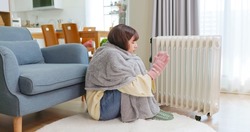 Side View Of Asian Woman Covering Blanket Wearing Gloves And Wool Hosiery Feel Cold Using Radiator Heater To Warm Up Sitting On Floor At Home In Living Room During Winter