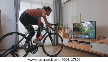 side view of Asian woman cyclist is exercising in the house by cycling on trainer and play online bike games with TV