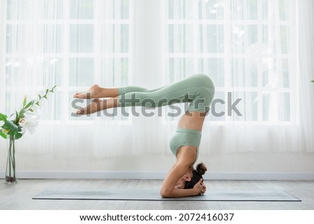 Side view of Asian woman doing Yoga exercise in front of windows,Yoga HandStand pose or Pincha Mayurasana,Calm of healthy young woman breathing and meditation yoga at home