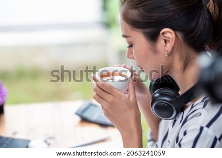 Side view of Asian woman in headphones sitting and relaxing while drinking coffee.