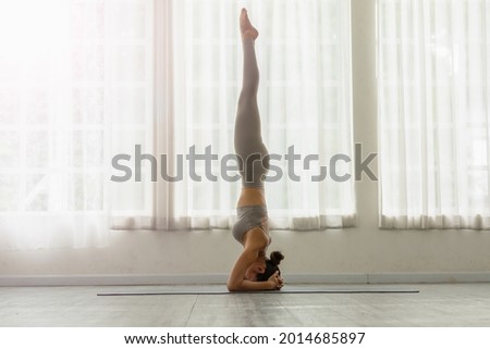 Side view of Asian woman wearing green sportwear doing Yoga Balance exercise in front of windows,Yoga HandStand pose or Pincha Mayurasana,Calm of healthy woman breathing and meditation yoga at home
