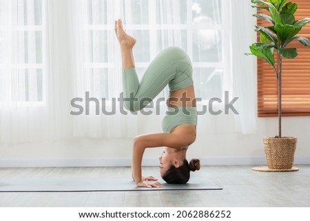 Side view of Asian woman wearing green sportwear doing Yoga exercise in front of windows.Yoga HandStand pose or Pincha Mayurasana.Calm of healthy young woman breathing and meditation yoga at home