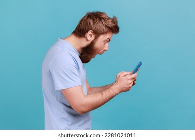 Side view of astonished surprised young adult bearded man standing, using smartphone and watching video with amazed face and open mouth. Indoor studio shot isolated on blue background.