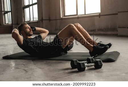 Side view of athletic male doing abdominal crunches while lying on mat and training in spacious shabby gym