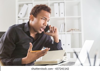 Side view of attractive african american businessman using notepad and laptop on office desktop while explaining something passionately over phone. Bookshelf with documents in the background