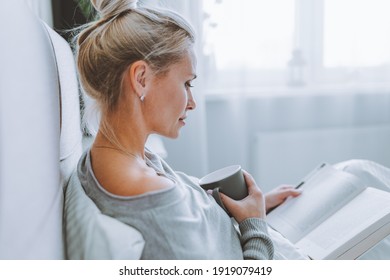 Side View Of Attractive Blonde Woman Sitting On Her Bed In Light Modern Bedroom With Grey Mug And Reading A Book.