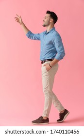 Side View Of An Attractive Businessman Wearing Blue Shirt Walking With One Hand In Pocket And Waving Excited On Pink Studio Background