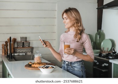 Side view of attractive Caucasian woman lady with blond wavy hair casual outfit standing in the modern kitchen having breakfast drink fresh smoothie juice holding using smart mobile cell phone - Powered by Shutterstock