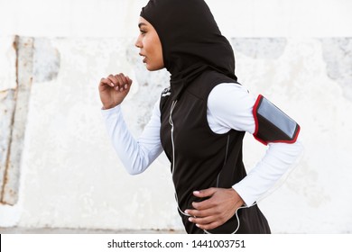 Side view of an attractive muslim sportswoman wearing hijab outdoors, listening to music with earphones, working out, running - Powered by Shutterstock