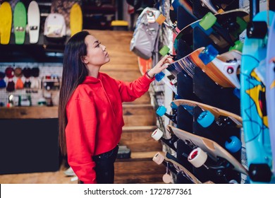 Side view of attractive teenage woman choosing longboard from store rack during Black Friday sales, beautiful Japanese hipster girl spending day for buying skateboard and practicing hobby skills - Powered by Shutterstock