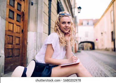 Side view of attractive wistful smiling woman with blond hair and in casual clothes writing in diary while sitting beside doorway on city street 