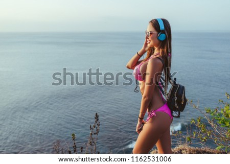side view of attractive woman in headphones and pink bikini looking at ocean from beach