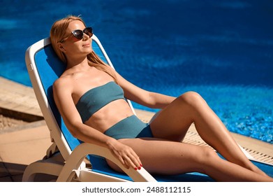 Side view of an attractive woman in a turquoise swimming costume lounging poolside. Woman enjoys relaxing by the pool with blue water. A girl relaxing under the tropical sun. Holiday concept. - Powered by Shutterstock